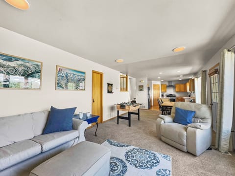 A living room with a gray sofa, white armchair, and a foosball table. The room leads into an open kitchen with wooden cabinets and white countertops. The walls have framed pictures.