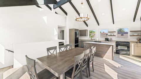 - Bright dining and kitchen area with open concept style  
- Wooden table with seating for group meals together  
- Modern chandelier and vaulted ceilings with beams