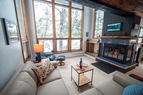 A cozy, sunlit living room featuring large lake-facing windows, a stone fireplace with a fire burning, and a comfortable gray sectional sofa.