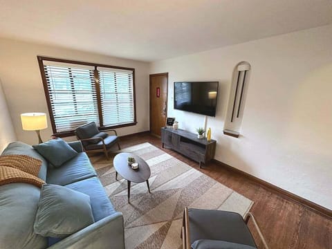 The living room features a blue sofa, two accent chairs, an oval coffee table, patterned rug, wall-mounted TV, media console, large windows, and front entry door.