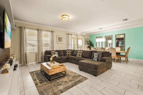 Open-plan living room with a spacious sectional, patterned pillows, and an adjacent dining area framed by bright windows and a turquoise accent wall.