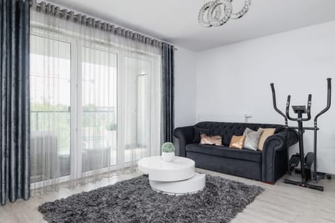 A modern living room with a sleek black sofa, a white coffee table, and floor-to-ceiling windows, creating a bright and stylish atmosphere.

