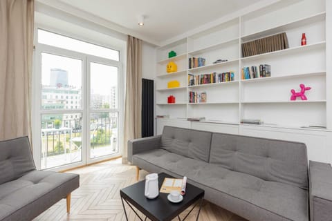 A bright living room with a grey sofa, modern shelves, and large windows offering a view of the city skyline.
