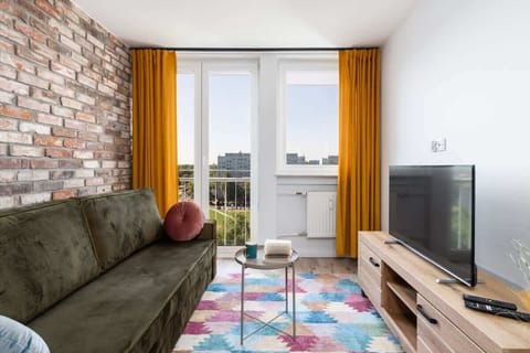 A cozy living area with a green sofa, colorful rug, bright yellow curtains, and a TV on a wooden stand.


