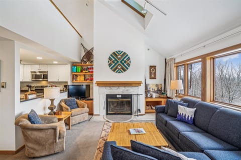 Bright, open living space with soaring ceilings, a wood-burning fireplace, and large windows that bring in natural light and scenic views.
