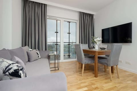 A modern living room with a gray sofa, a wall-mounted TV, and a dining area by the window, creating a bright and functional space.

