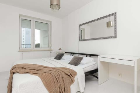 A bright bedroom with a white double bed, a large mirror above the bed, and a small desk in the corner, creating a functional and comfortable space.


