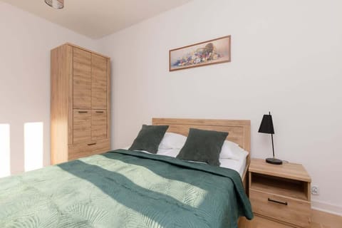 A cozy bedroom with a double bed covered in emerald green bedding, a wooden bedside table, and soft natural light streaming in.

