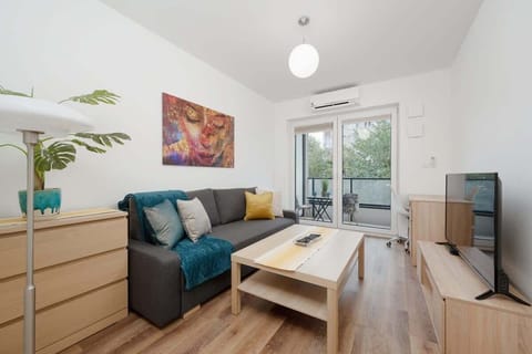 A bright living room with a gray sofa, yellow pillows, a wooden coffee table, and a large window letting in natural light.

