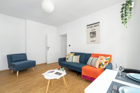 A bright living room contains a blue sofa, a small coffee table, and colorful cushions that add contrast to the white walls. Wooden flooring creates a warm atmosphere.