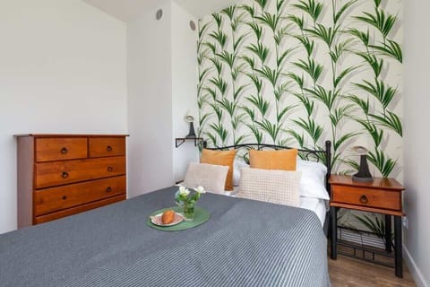 A cozy bedroom featuring a double bed with gray bedding, wooden furniture, and decorative wallpaper with a green leaf pattern.

