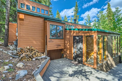A modern wooden cabin with large windows, set in a forested area. A stack of firewood is visible near the entrance. The scene is sunny with a blue sky and scattered clouds.
