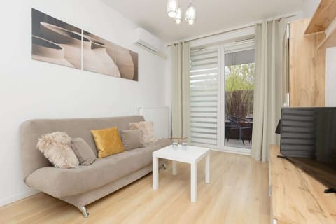 A bright living room featuring a beige sofa, a white coffee table, and large windows with light curtains, creating a cozy and inviting atmosphere.
