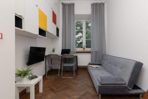 A bright living area with white walls, a gray sofa, colorful wall panels, and a wooden side table, complemented by gray curtains.

