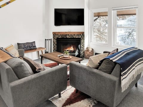 A warm and inviting living room featuring comfortable seating, a wood fireplace, wall-mounted TV, and large windows with snowy views.