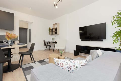 A bright living room with a gray sofa, wall-mounted TV, and a dining table. Decor elements add warmth to the modern space.
