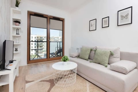 A bright living room with a gray sofa, green cushions, and a round coffee table. Large windows provide ample natural light.
