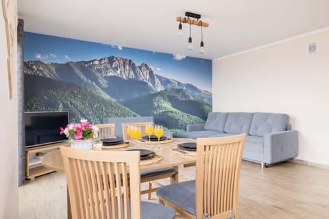 A bright dining and living area with a wooden table, chairs, and a mural of mountain scenery on the wall, creating a cozy and natural ambiance.
