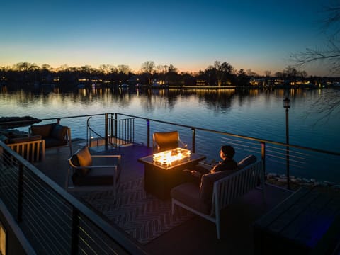 Rooftop deck fire pit lounge at twilight on the Magothy River, with a guest enjoying the warm glow as the lights of waterfront homes reflect across the calm water.