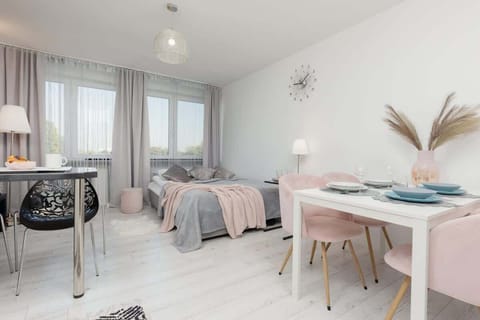 A bright living and dining area with large windows, light wooden flooring, a pastel pink dining set, and modern decor.
