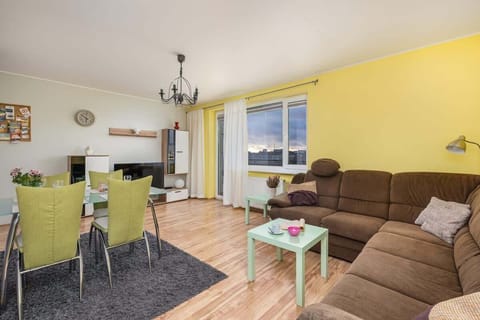 A bright living-dining area with yellow walls, a brown corner sofa, and a dining table with green chairs, creating a cozy and cheerful atmosphere.
