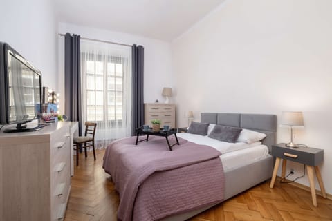 A cozy bedroom featuring a double bed with purple bedding, a bedside lamp, and a large window with curtains.
