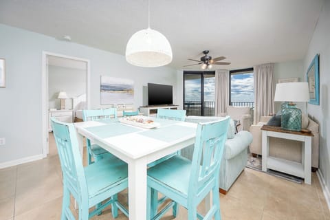 Bright dining area with turquoise chairs, a white table, and a view of the beach through large windows in a coastal space