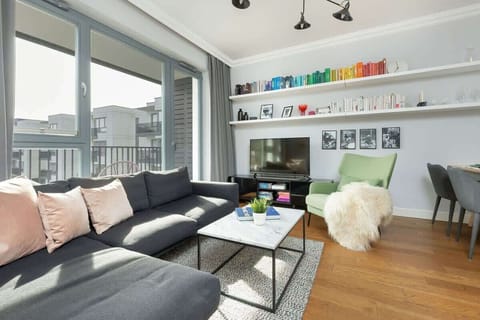 A bright living area with a grey corner sofa, colorful books on display, and large windows overlooking the city.
