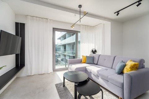 A bright living room with a gray sofa, a round coffee table, and a wall-mounted TV. Sheer curtains let in natural light.
