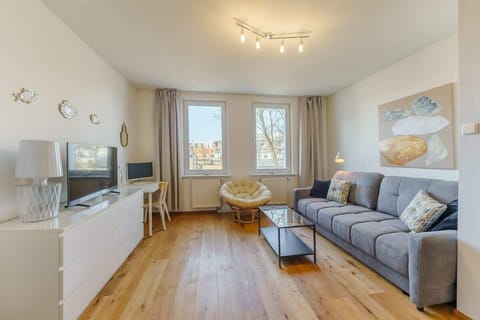 A spacious living room with a gray sofa, a white TV stand, and large windows allowing natural light to fill the space.
