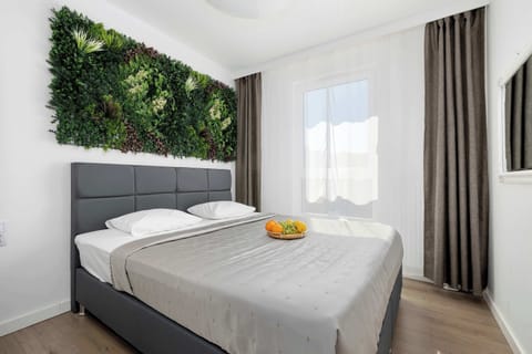 A bright bedroom with a double bed, white bedding, curtains, and a decorative green plant wall above the headboard.