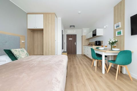 A bright bedroom with a double bed, soft pink bedding, and a wooden wardrobe. A small dining area with a round table and turquoise chairs is visible in the background.