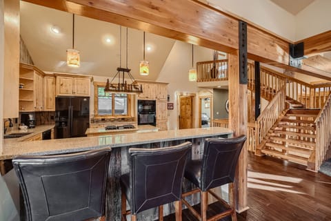 A modern kitchen with wooden cabinets and a large island. Three black barstools line the island, and a wooden staircase is visible in the background. Hanging lights illuminate the kitchen.