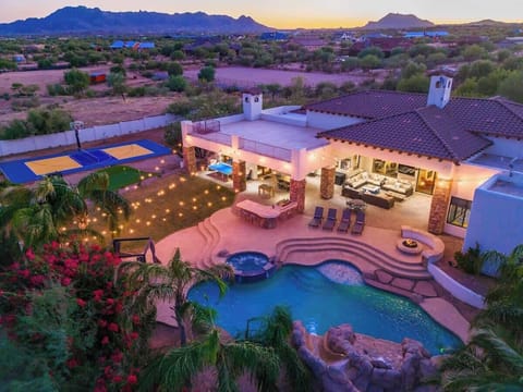 Stunning aerial view captures a luxurious desert estate at dusk. The property features a resort-style pool, hot tub, sports court, and outdoor entertaining areas, all set against dramatic mountain vistas and sprawling desert landscape.