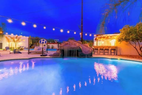 Twilight transforms the backyard into a glowing retreat. String lights reflect off the pool, while a rock waterfall and outdoor bar set the stage for unforgettable evenings under the stars.