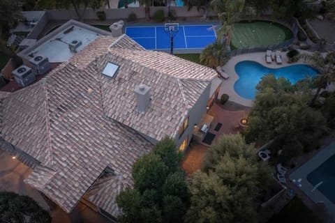 Aerial overview of the rental property featuring a tiled-roof home, pristine swimming pool, full-size tennis court, and putting green surrounded by lush landscaping in a peaceful residential neighborhood.