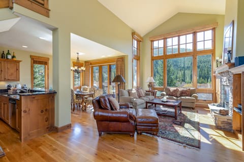 A spacious living room with large windows, wooden flooring, and a mix of leather and fabric furniture. The room features a mountain view, a stone fireplace, and an adjoining dining area and kitchen.