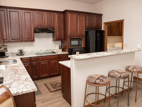 A kitchen with dark wooden cabinets, granite countertops, a black refrigerator, oven and microwave, and a breakfast bar with three cushioned stools.