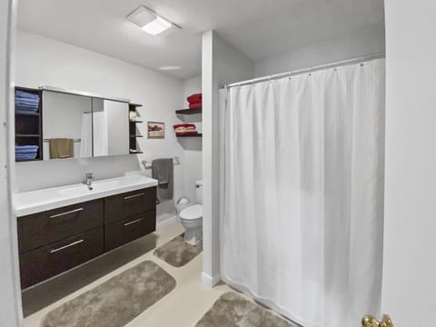 A modern bathroom with a large mirror, sink cabinet, toilet, and shower with a white curtain. The room features a shelf with folded towels and is accented by neutral-colored decor.