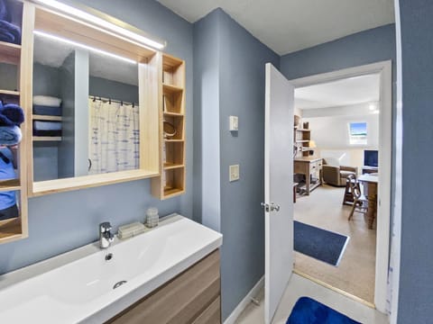 A bathroom with a sink, mirror with shelves, and partially visible shower curtain. The door is open, leading to a room with a couch, chair, and desk. Blue walls and rugs accent the space.