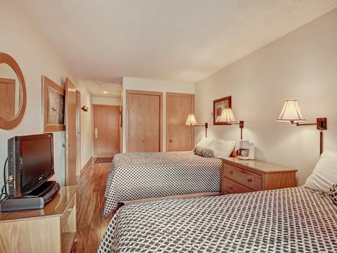 A hotel room with two double beds, a small flat-screen TV on a dresser, wall-mounted lamps, and wooden furnishings. The room has white walls, patterned bed covers, and a wooden floor.
