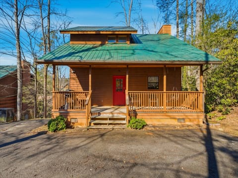 Charming log cabin with green metal roof, welcoming red door, and wraparound porch nestled among tall trees.