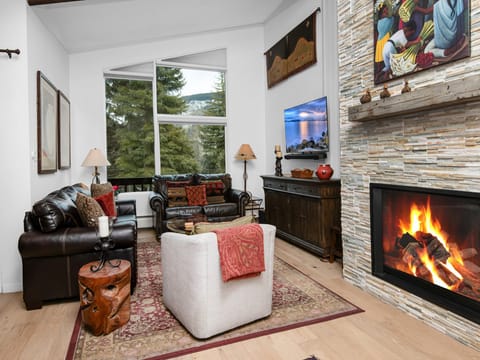 Cozy up by your crackling fireplace while enjoying forest views through expansive windows in this inviting living room.