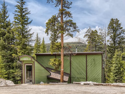 A green wooden cabin with the address "683" is nestled in a forested area, with a tall tree growing through its deck. Snow-capped mountains are visible in the background.