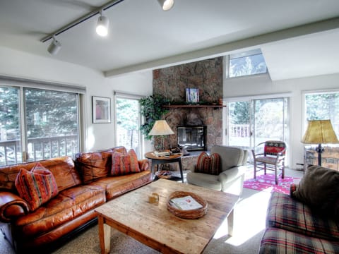 A cozy living room with large windows, brown leather sofa, a wooden coffee table, armchairs, and a stone fireplace. Sunlight streams in, and there is a view of trees outside.
