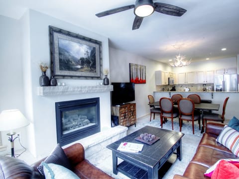 Modern living room and dining area featuring a fireplace, ceiling fan, sectional sofa, coffee table, and dining set. An open kitchen with bar seating is visible in the background.
