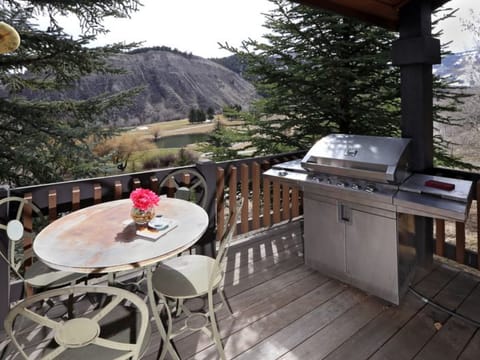 A wooden deck with a gas grill and a round metal table with three chairs and a vase of flowers, overlooking a hilly landscape with trees.