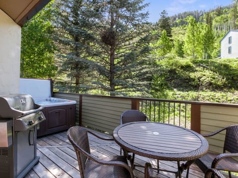 A wooden deck with a round table and four chairs, a gas grill, and a hot tub, overlooking a wooded hillside on a sunny day.
