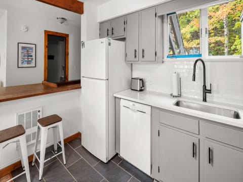 A modern kitchen with gray cabinets, a white refrigerator, a white dishwasher, a stainless steel sink, a toaster, and two white stools at a wooden breakfast bar. A window provides natural light.