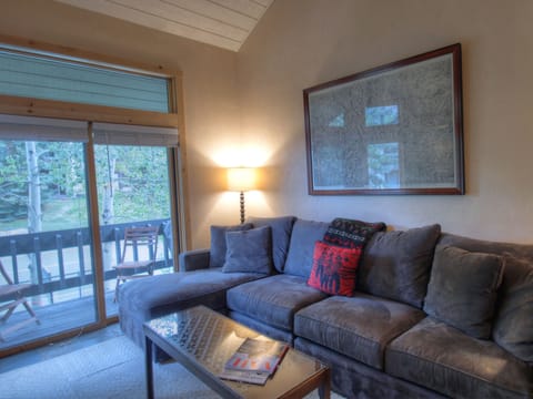 A living room with a gray sectional sofa, a red accent pillow, a coffee table with magazines, a floor lamp, a framed picture on the wall, and glass doors leading to a balcony with outdoor chairs.